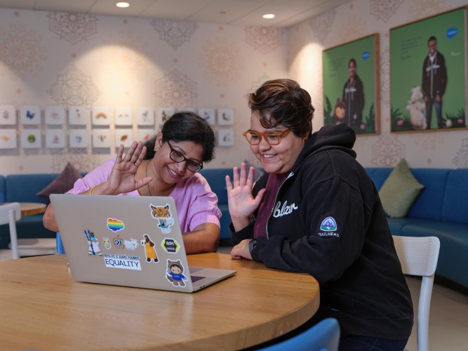 two women sit at a table in a modern office setting and wave at a computer