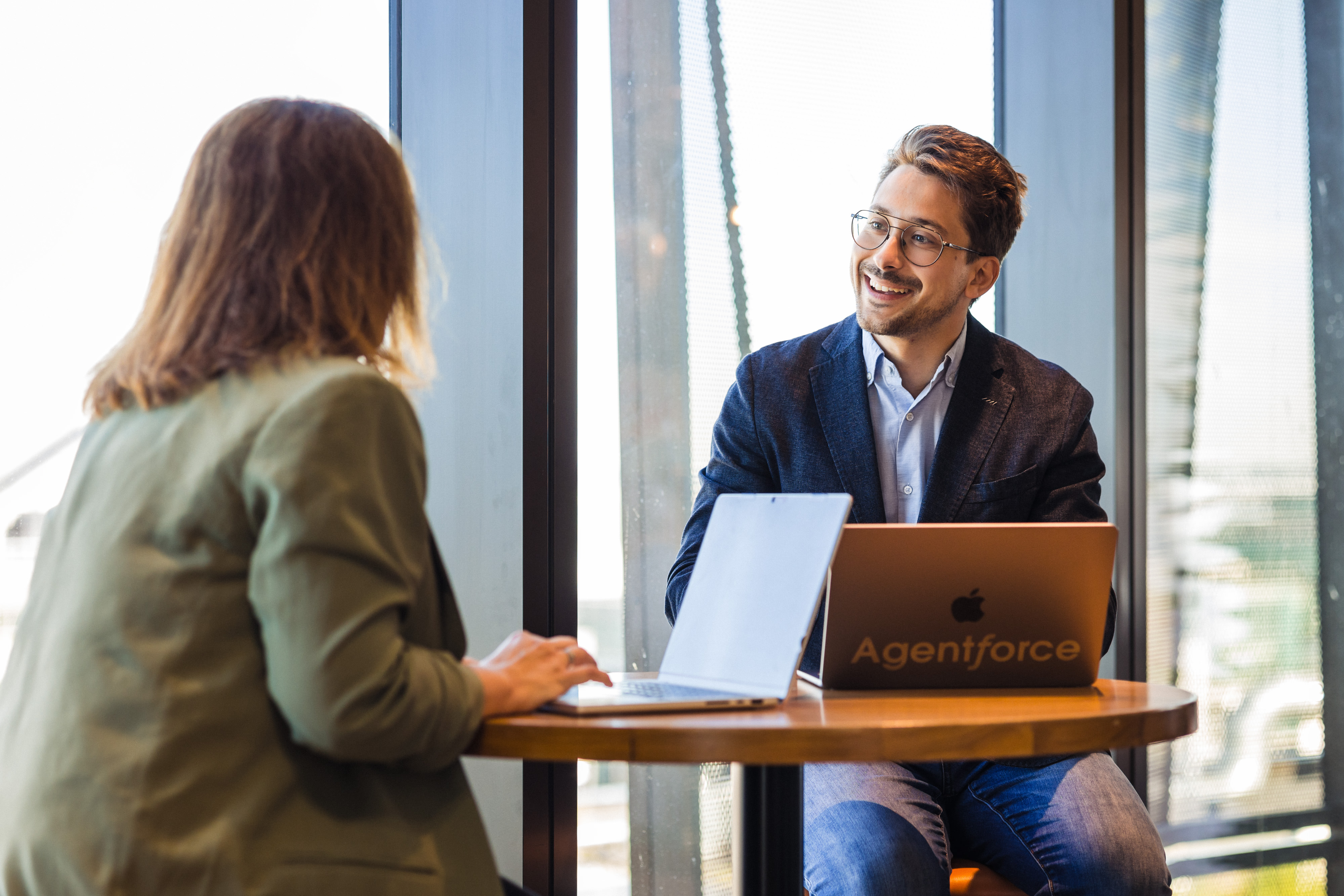 One person smiles at the camera while working on a laptop, another at the desk smiles while looking at the laptop.