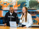 two employees stand together in a modern office space and collaborate around a computer