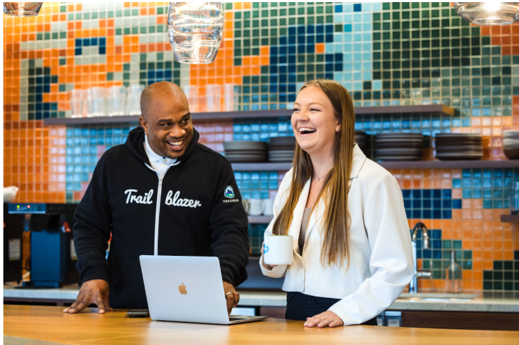 two employees stand together in a modern office space and collaborate around a computer