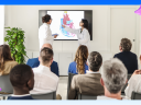 Two medical professionals in white lab coats present a colorful anatomical illustration of a human heart on a large display screen to a seated audience of approximately eight people in a bright, modern conference room.