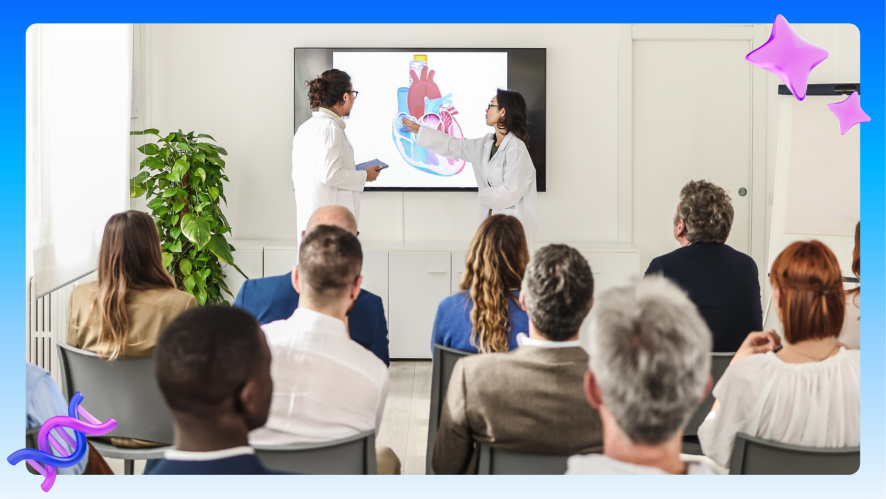 Two medical professionals in white lab coats present a colorful anatomical illustration of a human heart on a large display screen to a seated audience of approximately eight people in a bright, modern conference room.