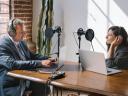 A man and woman recording a podcast in an office setting at a desk with microphones and equipment.