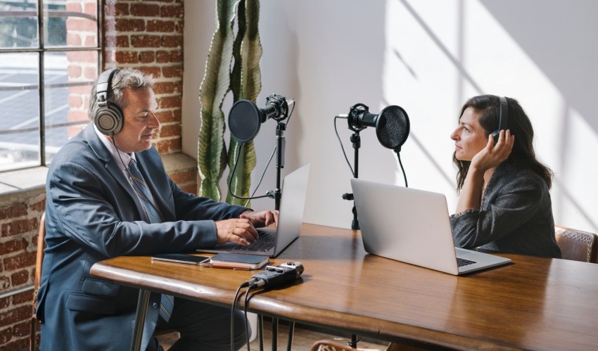 A man and woman recording a podcast in an office setting at a desk with microphones and equipment.