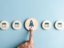 A line of wooden icons showing SaaS products and a finger pointing to the middle one on a blue background.