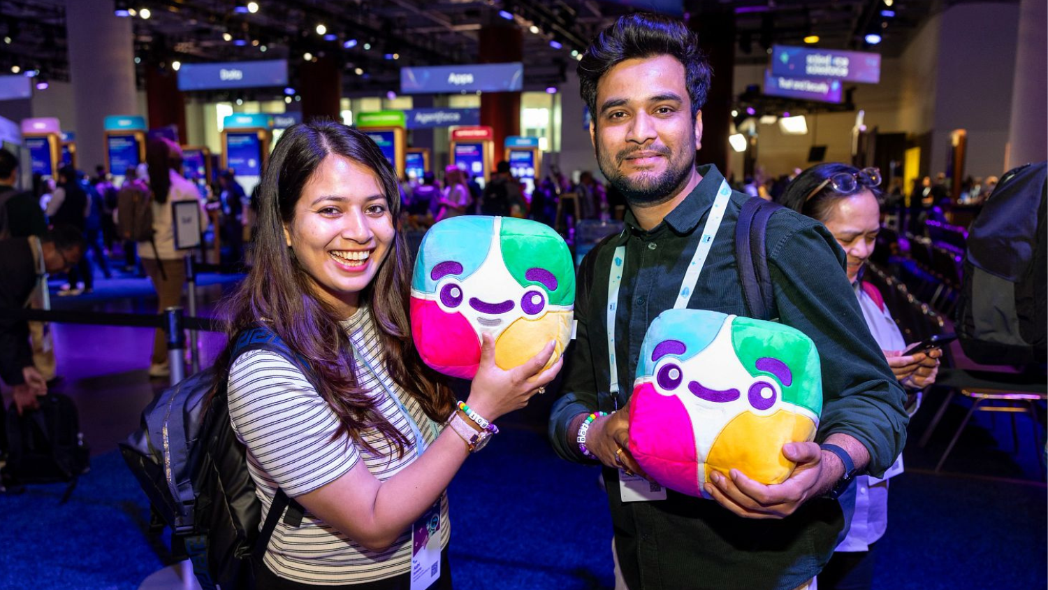 Two attendees at a Salesforce event smile while holding colorful mascot plush toys on a busy expo floor.