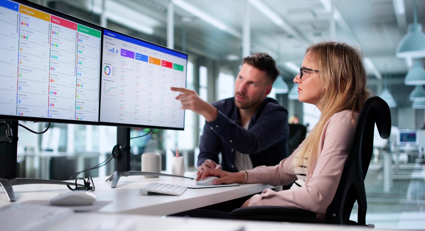 A man and woman sit at a desk looking at opportunity management software on two computer screens.