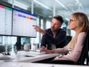 A man and woman sit at a desk looking at opportunity management software on two computer screens.