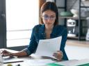 Business woman working with computer while consulting some invoices and documents in the kitchen at home.