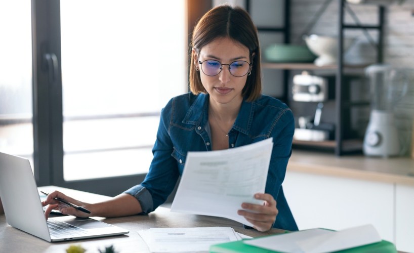 Business woman working with computer while consulting some invoices and documents in the kitchen at home.