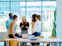 A small office with people talking around a desk in front of windows.