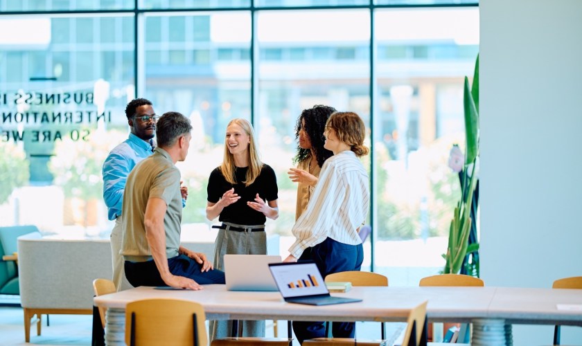 A small office with people talking around a desk in front of windows.