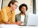Two colleagues have pleasant conversation sitting side by side in modern open space office in front of laptop, smiling and enjoying friendly working atmosphere, brainstorming together, solving tasks.
