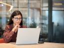 Businesswoman wearing glasses reviewing information on a laptop at a wooden desk in a modern office