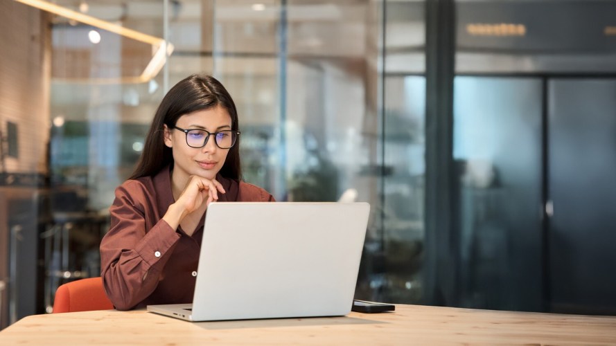 Businesswoman wearing glasses reviewing information on a laptop at a wooden desk in a modern office