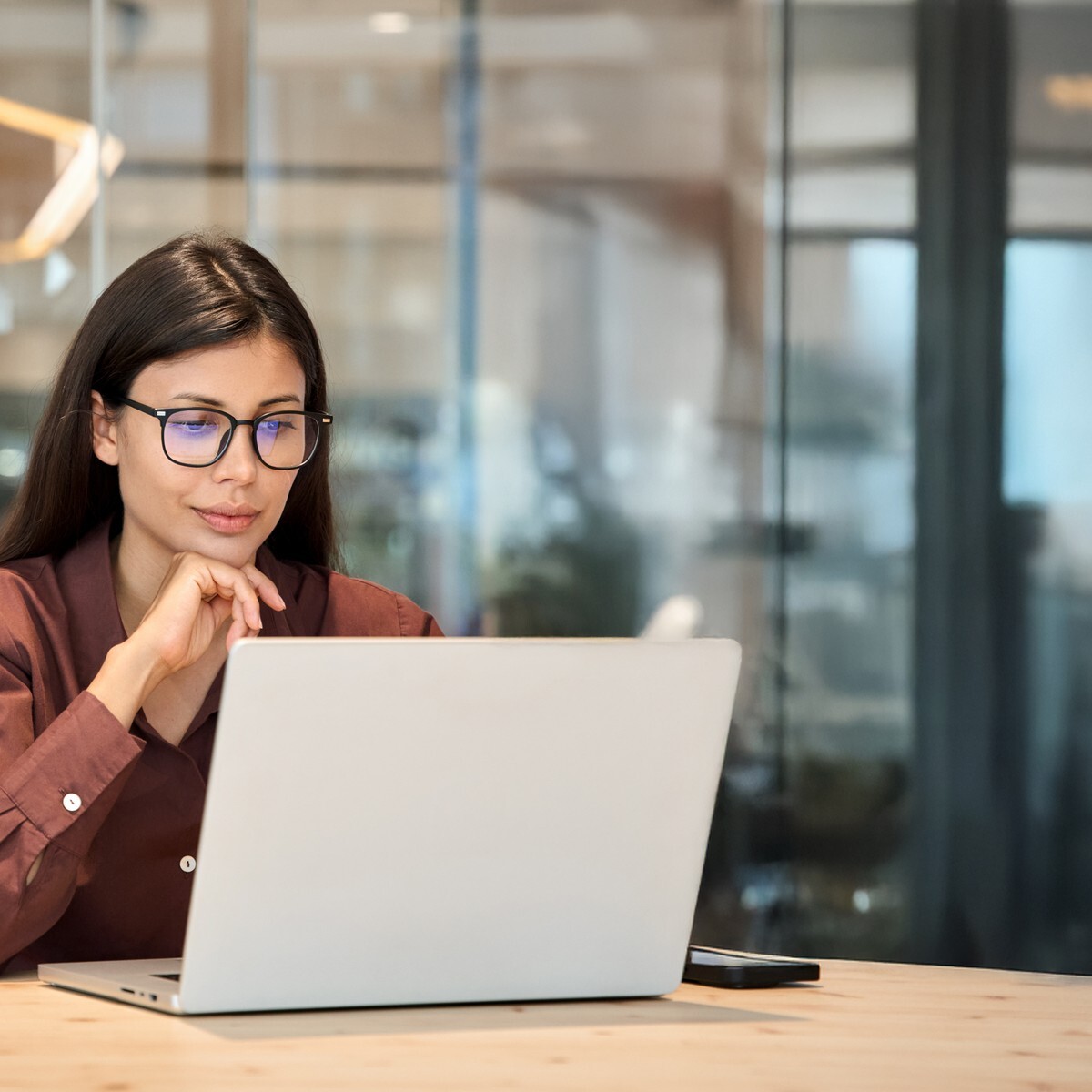 Businesswoman wearing glasses reviewing information on a laptop at a wooden desk in a modern office