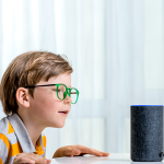 Garoto interagindo com uma caixa de som, que possui assistente virtual, que está sobre a mesa