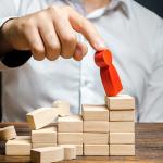 Homem derruba pequeno boneco vermelho de madeira do topo de uma pirâmide, representando a carreira corporativa.