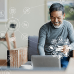 Homem sentado em uma poltrona na sala mexendo em um notebook que está em uma mesa à sua frente. Em volta do computador há desenhos de ícones fazendo alusão à navegação na internet, como carrinho de compras, arroba, fones de ouvido, e-mail.