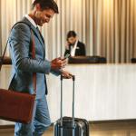 Homem de terno, na recepção do hotel, puxando sua mala e interagindo com o celular.