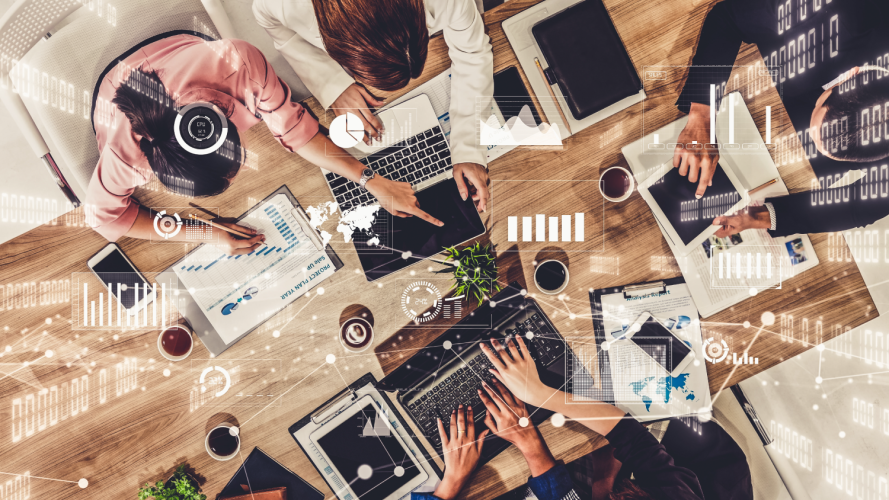 Uma vista aérea de uma equipe colaborando em uma mesa de madeira, rodeada de laptops, tablets, gráficos e documentos analíticos. As pessoas estão revisando e discutindo informações financeiras e de projetos, apontando para telas e documentos impressos. Elementos digitais, como gráficos de barras, indicadores de desempenho e mapas, estão sobrepostos na imagem, simbolizando o uso de tecnologia e análise de dados em tempo real para a tomada de decisões. A atmosfera é dinâmica e colaborativa, representando um ambiente de trabalho moderno e focado em análise estratégica.
