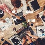 Uma vista aérea de uma equipe colaborando em uma mesa de madeira, rodeada de laptops, tablets, gráficos e documentos analíticos. As pessoas estão revisando e discutindo informações financeiras e de projetos, apontando para telas e documentos impressos. Elementos digitais, como gráficos de barras, indicadores de desempenho e mapas, estão sobrepostos na imagem, simbolizando o uso de tecnologia e análise de dados em tempo real para a tomada de decisões. A atmosfera é dinâmica e colaborativa, representando um ambiente de trabalho moderno e focado em análise estratégica.