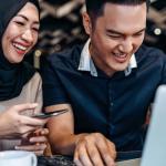 Um casal jovem sorri enquanto colabora em um laptop em um ambiente moderno e descontraído. A mulher, que usa um hijab preto e uma blusa bege, segura um celular, enquanto o homem, vestindo uma camisa azul, parece concentrado no teclado do laptop. A cena reflete trabalho em equipe, conexão digital e uma atmosfera amigável, provavelmente em um café ou espaço de coworking. Ao fundo, luzes difusas adicionam um toque acolhedor ao ambiente. Ideal para ilustrar temas de empreendedorismo, tecnologia ou trabalho remoto.