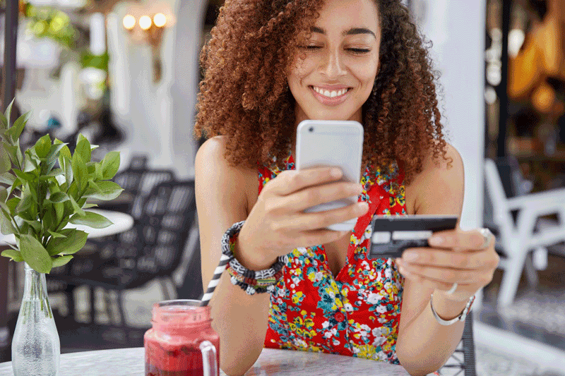 Eine Frau sitzt in einem Restaurant und hat ihr Handy sowie eine Kreditkarte in der Hand, um auf einem der Online-Marktplätze einzukaufen