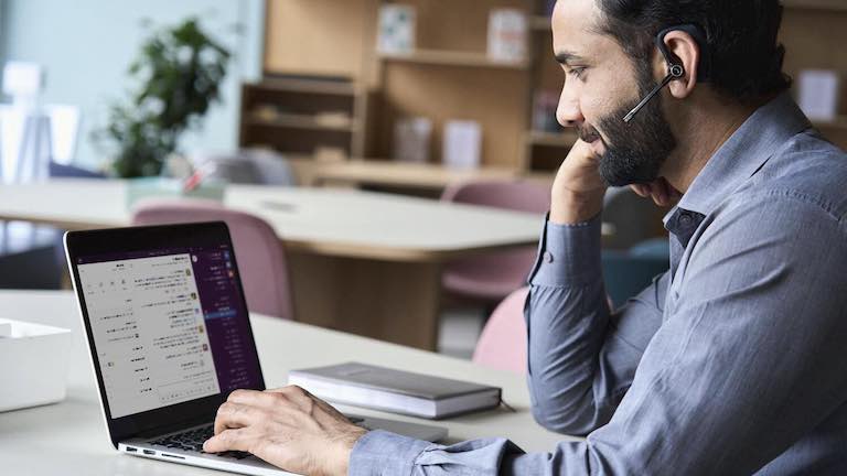Un homme barbu, portant un casque et assis à son bureau, utilise Slack sur un ordinateur portable