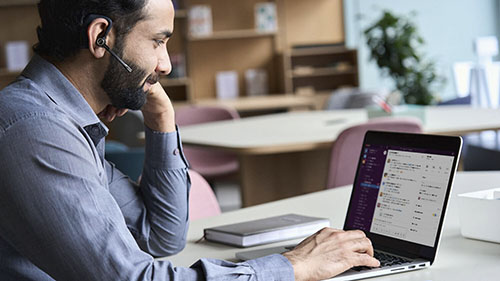Man med skägg och headset använder Slack på bärbar dator vid skrivbord