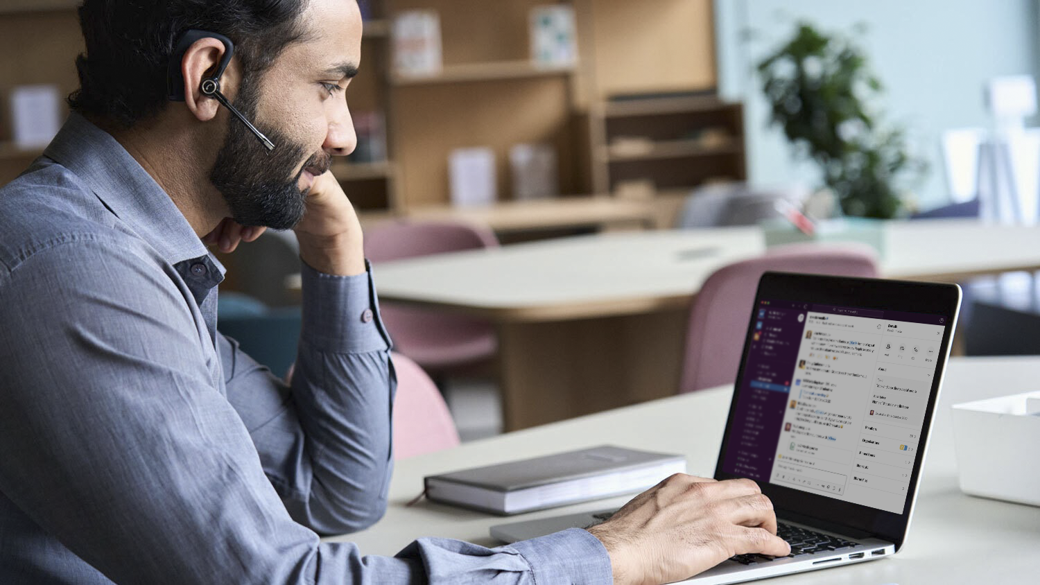 A man with a beard and headset at a deck uses slack on a laptop 