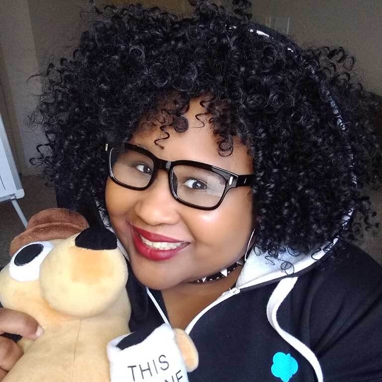A woman with curly dark hair, wearing dark-rimmed glasses, holds up a plush doll of a dog.