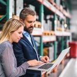 eine Frau und ein Mann stehen in einer Lagerhalle und schauen auf einen Laptop