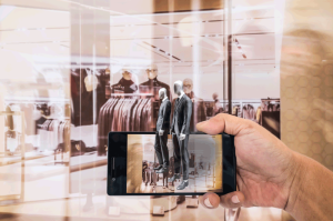 Eine Person hält ein Smartphone in der Hand und im Hintergrund ist ein Geschäft zu sehen. Die Grenzen zwischen Offline und Offline Handel verbinden sich.