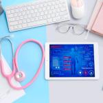 Auf einem Tisch liegt eine Tastatur, ein iPad, ein Notizbuch, ein Stethoskop, eine Brille, Papier etc.