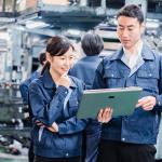 Two workers in a factory — a woman and a man, both wearing dark blue jumpsuits — look at plans on a tablet. / best field service management software