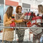Three women stand in a mall holding wrapped boxes. Holiday shopping predictions say it’s likely they paid for them with credit cards.