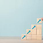 A hand places a wooden block with an upward-pointing arrow onto a staircase of similar blocks.