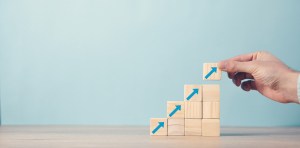 A hand places a wooden block with an upward-pointing arrow onto a staircase of similar blocks.