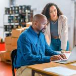 Two coworkers, a man and a woman, sit at a table in a warehouse, looking at a laptop / large language models