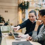 Two young businesswomen sitting at a desk and looking at a computer. The woman on the left is pointing at the computer screen.
