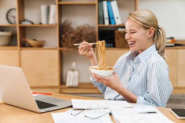 Image of a smiling woman, sitting at a desk with a laptop and papers, very excited to eat a bowl of spaghetti with chopsticks