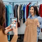 Two women shop for clothing in a store. One woman is taking a photo of her friend, who is holding up a light brown dress. / referral marketing