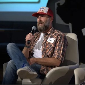 Photo of the author named Scott Leese sitting in a chair with a mic in his hand during a public event.