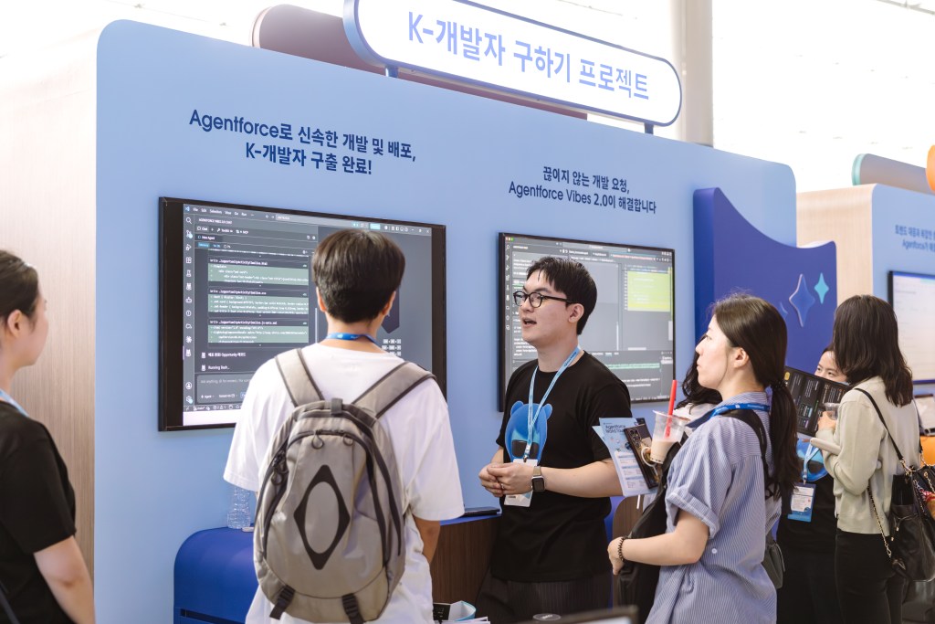 세일즈포스 Agentforce World Tour Korea 컨퍼런스의 개발자 부스 전경과 모니터에 띄워진 바이브 코딩(Vibe Coding 2.0) 화면을 살펴보며 대화를 나누는 사람들