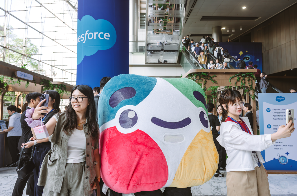 세일즈포스 Agentforce World Tour Korea 컨퍼런스 홀 내부에서 대형 브랜드 기둥과 Slackbot 캐릭터 인형을 배경으로 기념사진을 찍고 있는 방문객들