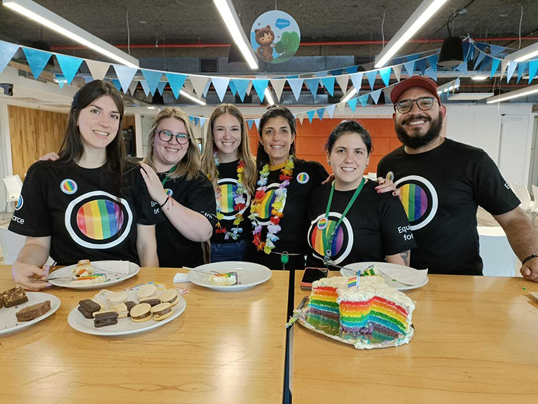Seis personas con camisetas con los colores de la bandera LGBTQ+, alrededor de una mesa con un pastel con los colores del arcoíris.