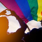 Silueta de personas, contra el sol, participando en una marcha, con una bandera LGBTQ+ encima.