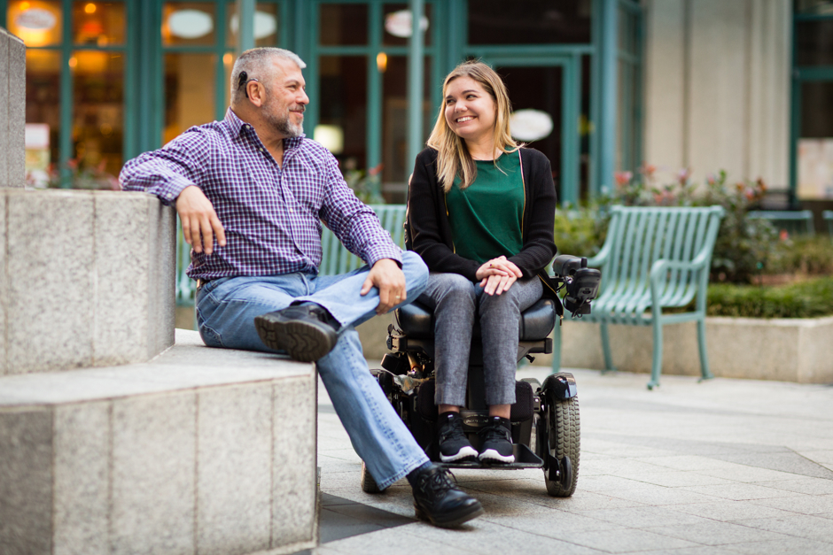 A man with a cochlear implant and a woman in a wheelchair are seated in an accessible outdoor patio area at one of Salesforce’s offices.