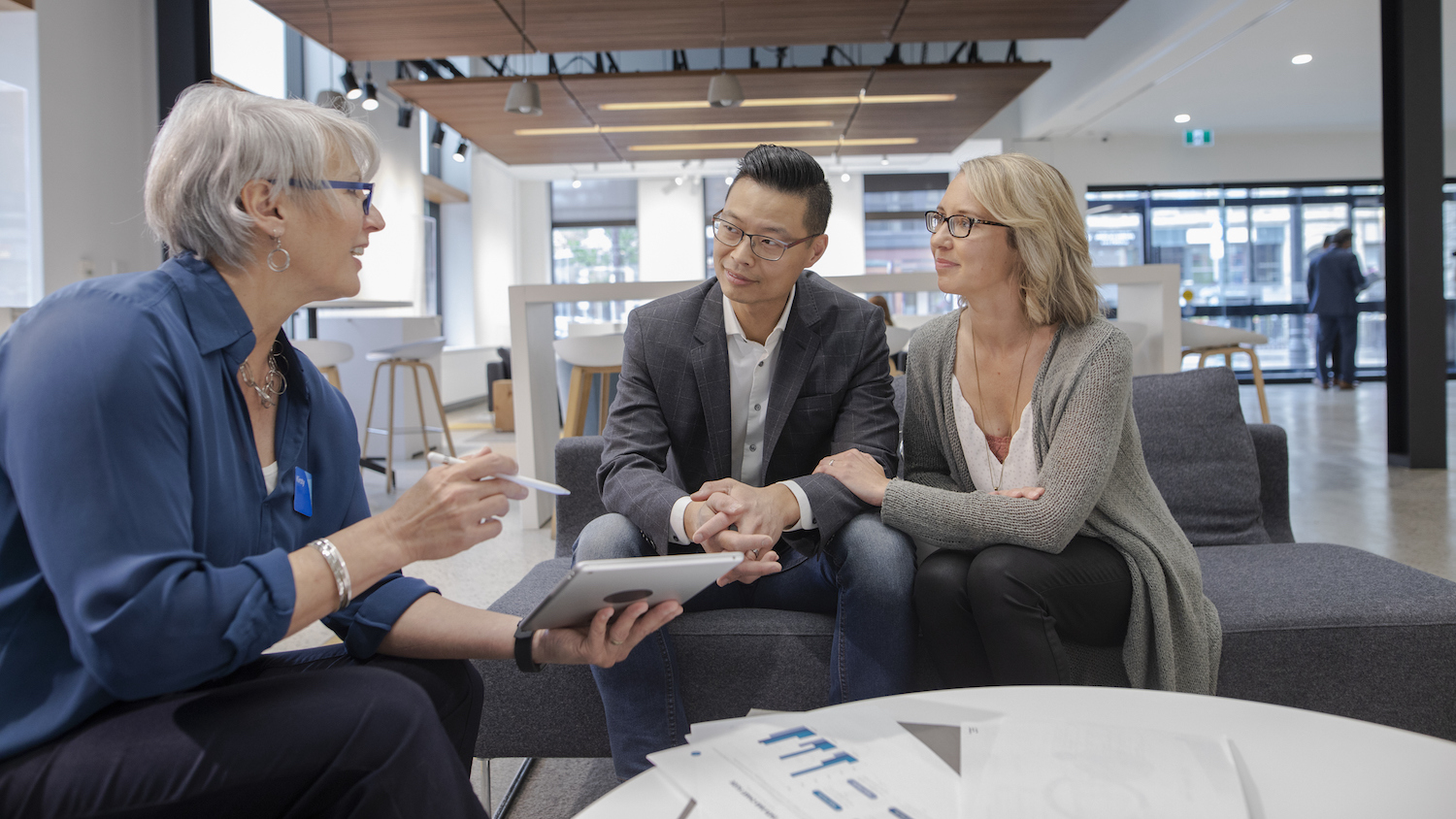 Photo of a female bank employee in a bank branch, sitting at a table and holding a tablet while helping a professional looking couple with customer service in banking